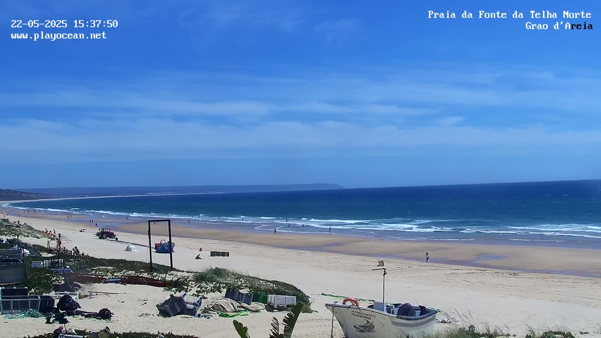 A sandy beach with a few people and a small boat is visible, under a mostly sunny sky with a blue ocean and small waves in the background.