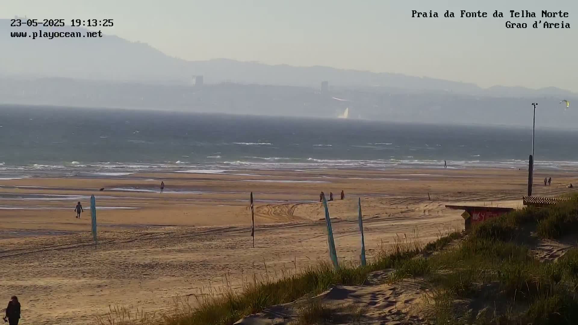 A hazy, sunny day at a beach shows a wide expanse of sandy shore with a few people scattered about, gentle waves lapping at the sand, and distant mountains visible on the horizon.