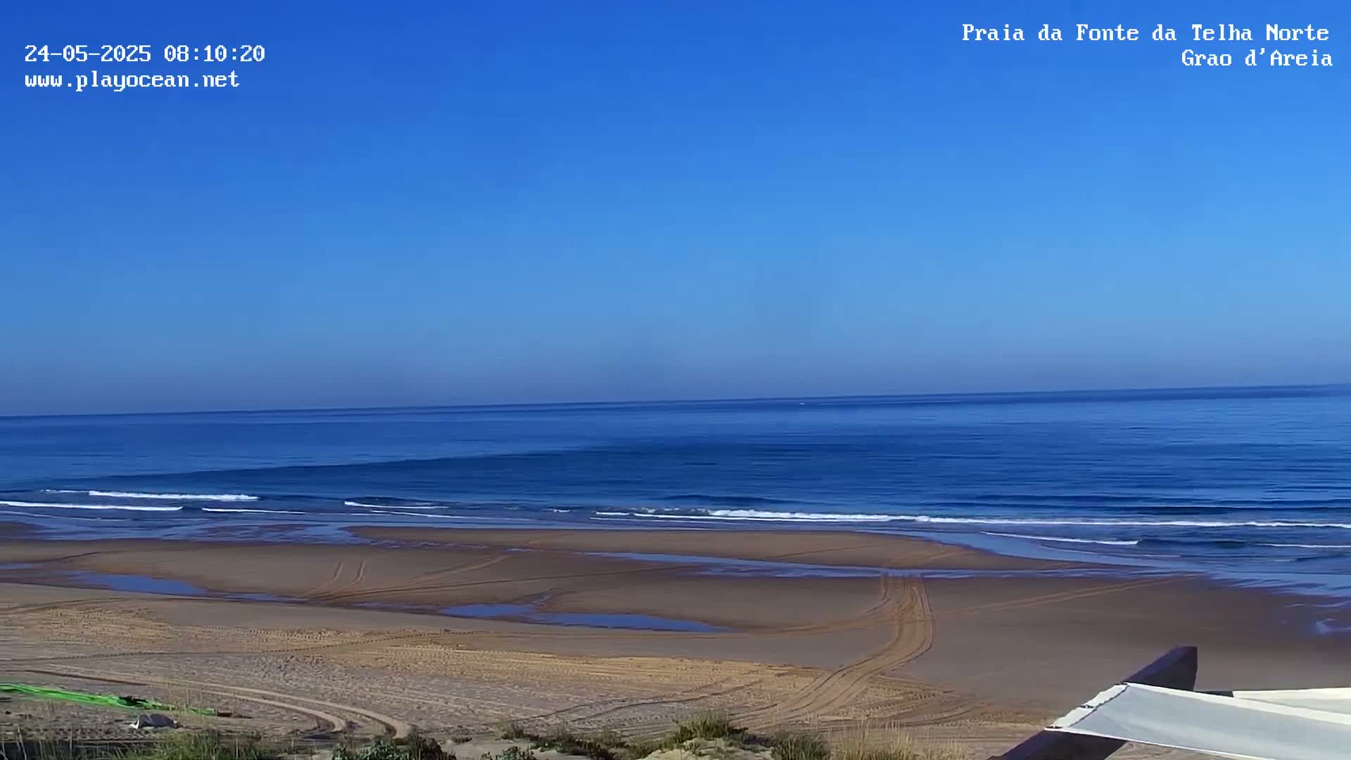 A sandy beach with tire tracks stretches to a calm, blue ocean under a clear blue sky.