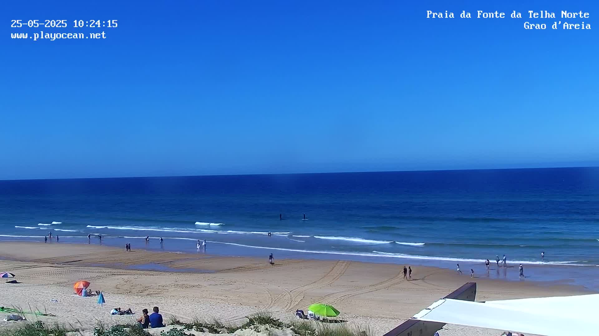 A sandy beach with a few people on it, under a clear blue sky, stretches towards a calm, blue ocean with small waves.