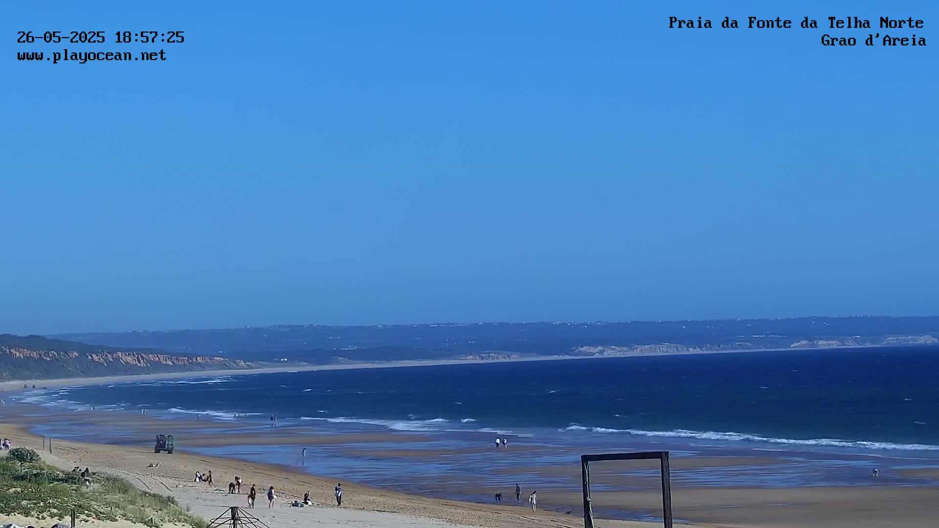 A sandy beach with a few people on it slopes gently down to a calm ocean under a clear blue sky.