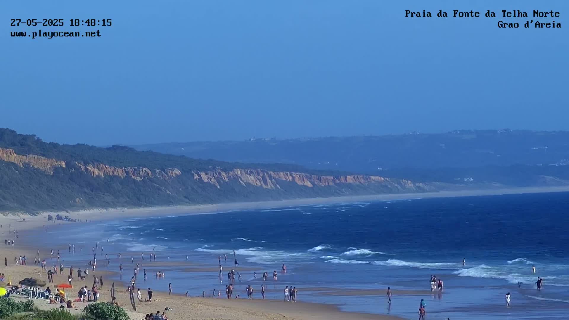 A sandy beach with many people on it is bordered by a line of cliffs under a clear blue sky, and waves are gently lapping the shore.