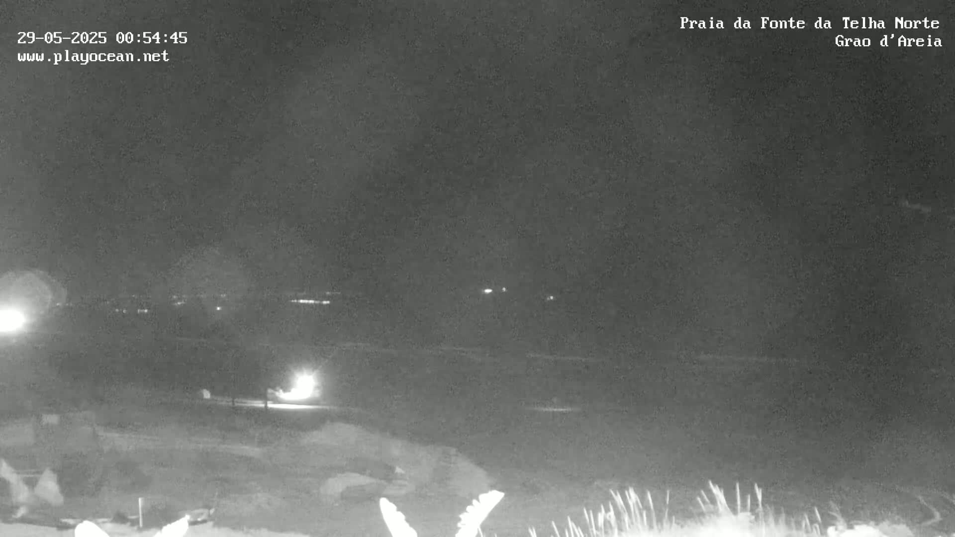 A nighttime, low-light view of a dark beach with a few distant lights visible on the horizon.
