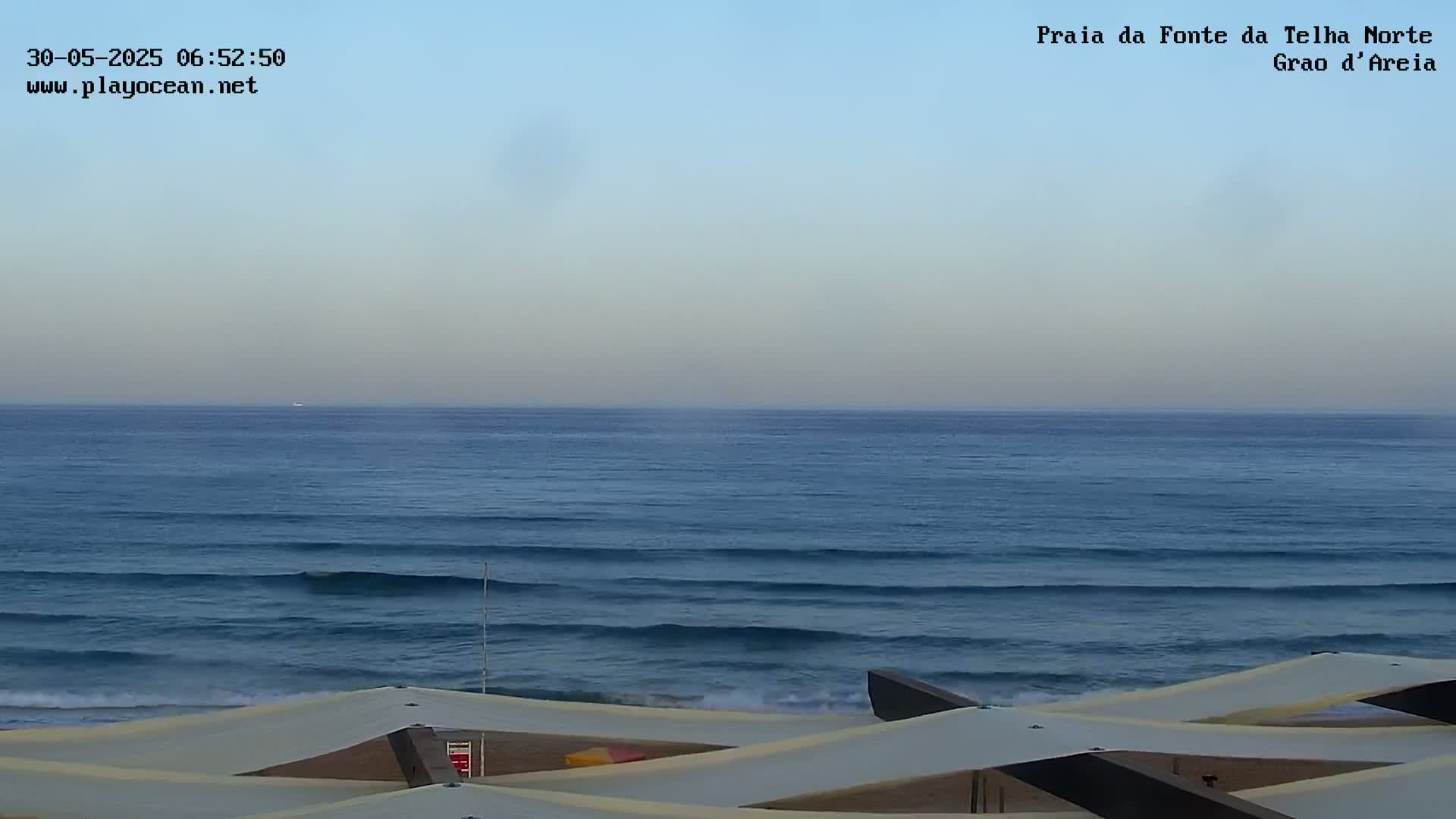 A calm ocean with small waves rolls onto a sandy beach under a clear, light-blue sky.