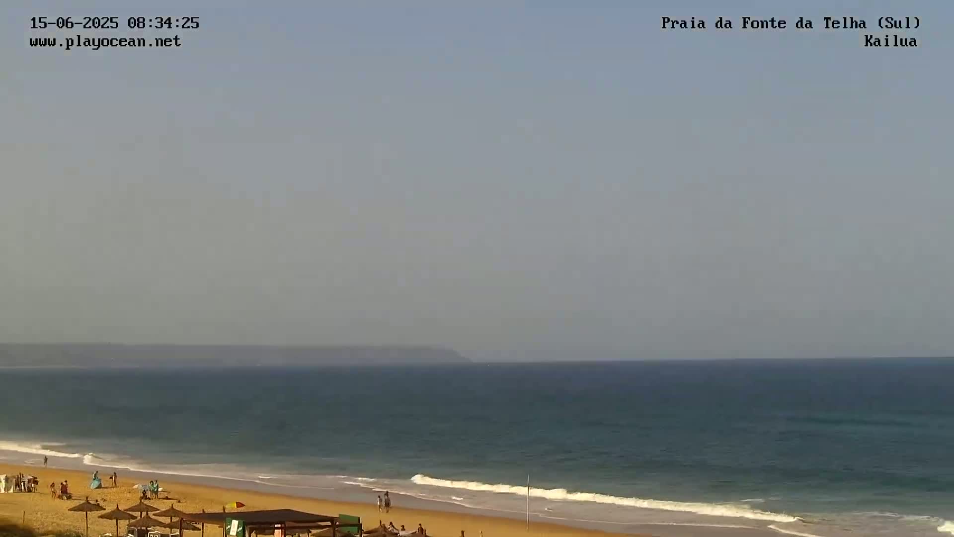 A hazy, sunny day at a beach with calm waves, showing a sandy shore with some people and umbrellas.