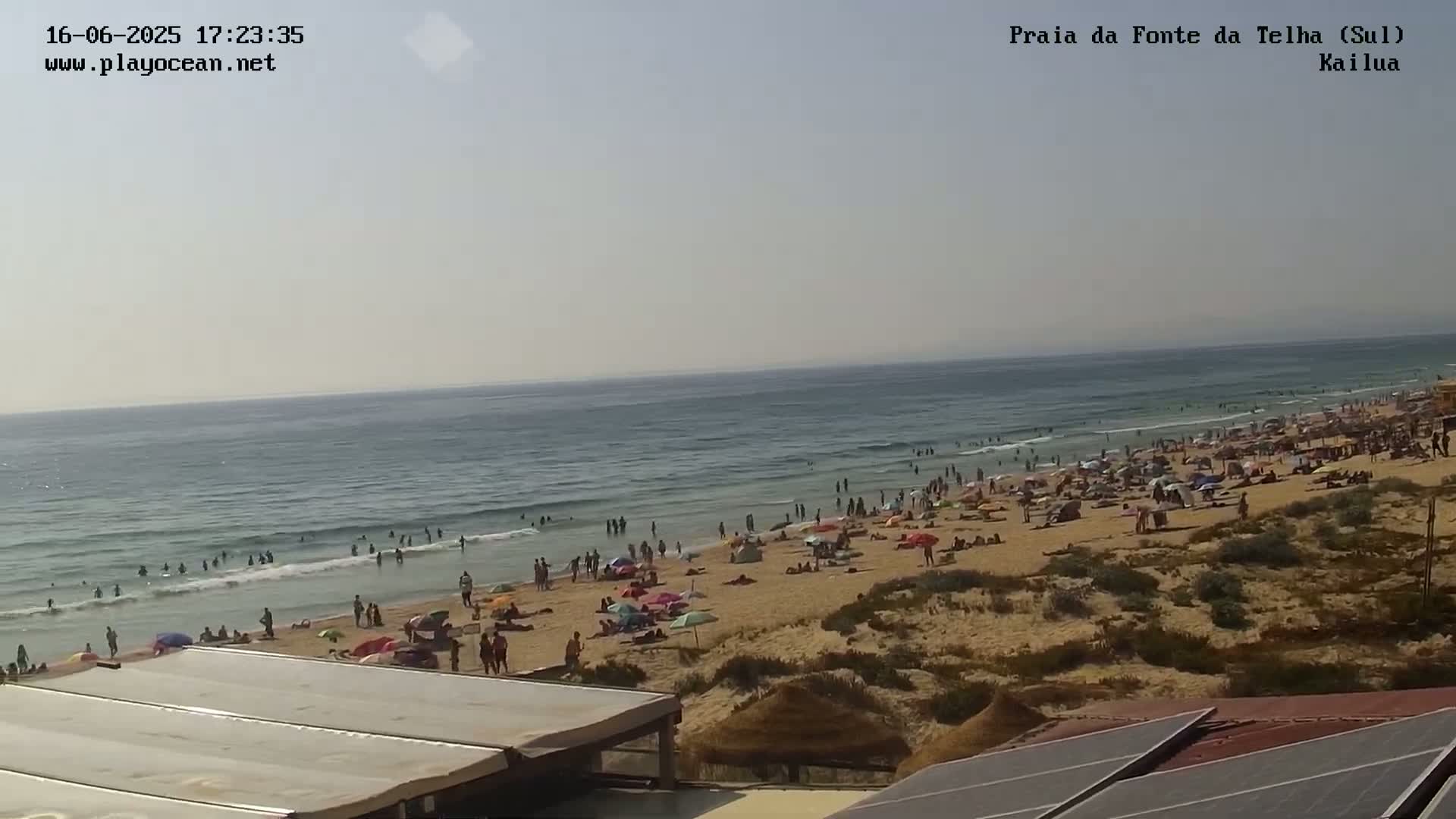 A crowded sandy beach with people swimming and sunbathing under a partly cloudy sky next to a calm ocean.