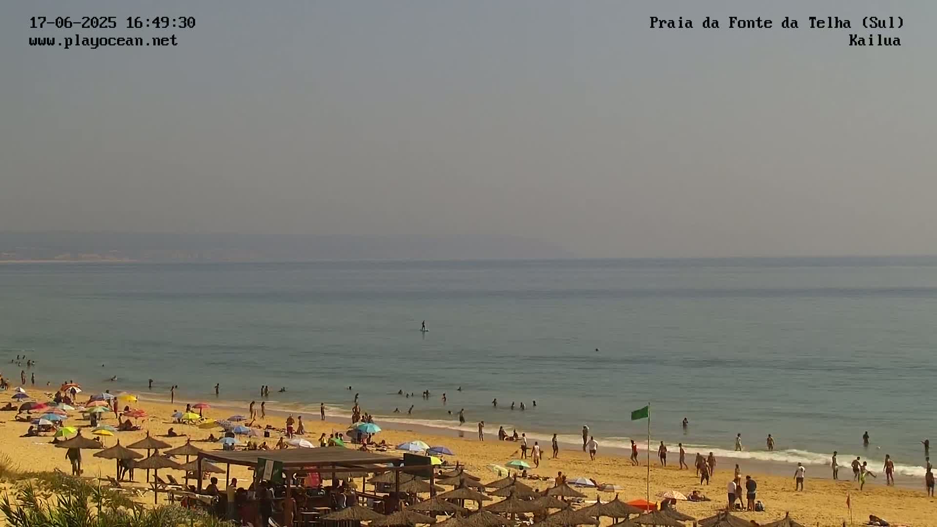 A sandy beach with many people and umbrellas under a hazy sky, and calm, light-blue ocean.