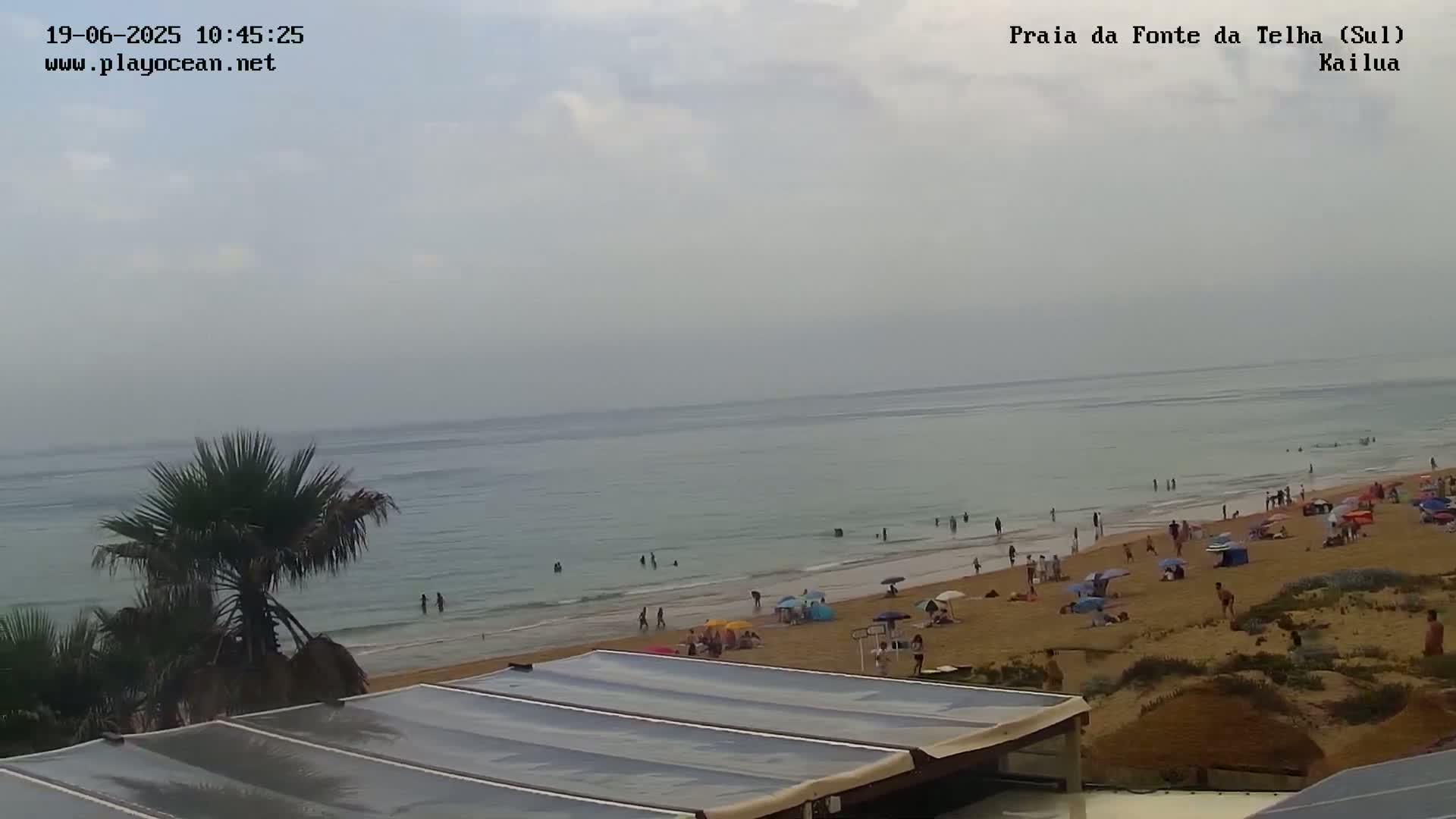 A sandy beach with numerous people and umbrellas under an overcast sky, features a calm ocean in the background.