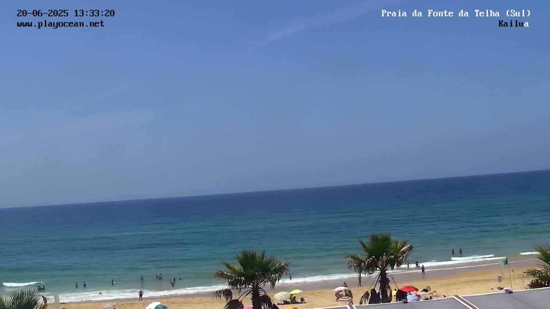 A sandy beach with palm trees and people swimming and sunbathing under a clear blue sky.
