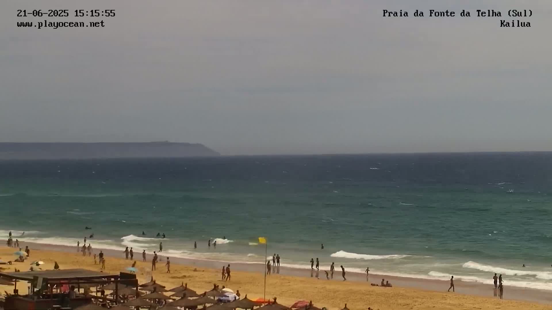 A sandy beach with people swimming and sunbathing, under an overcast sky, meets a teal ocean with small waves, and a distant landmass is visible on the horizon.
