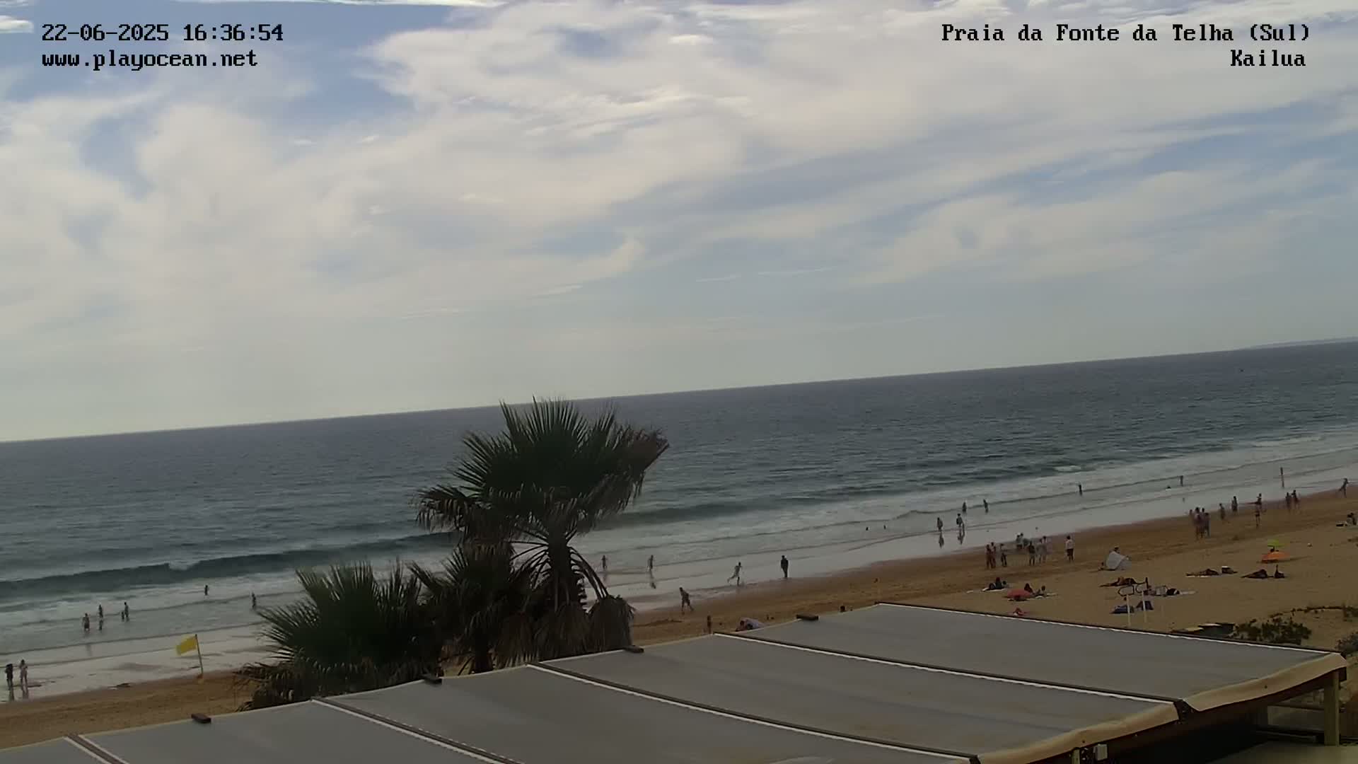 A partly cloudy day at a beach shows a calm ocean with people on the sand and in the water, palm trees in the foreground, and a section of a building's roof.