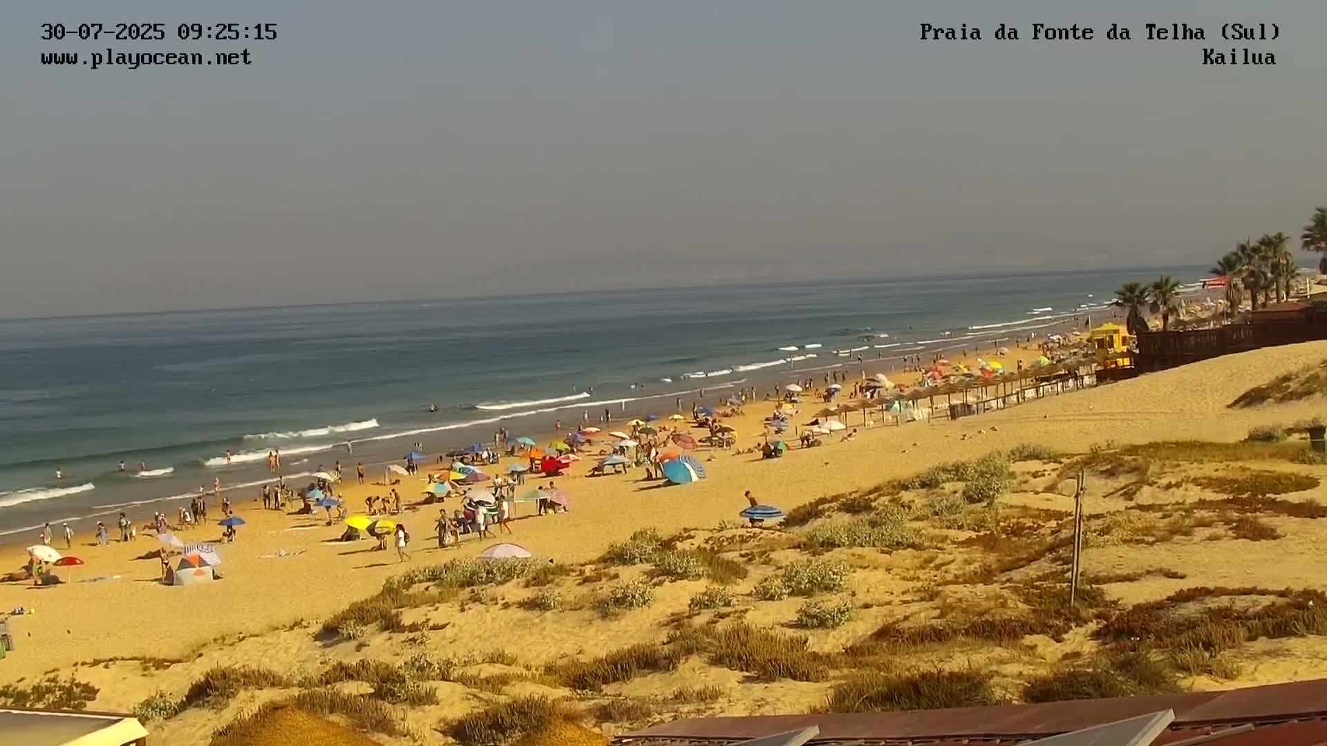 A sandy beach with many people under colorful umbrellas and some surfers in the ocean on a hazy day.