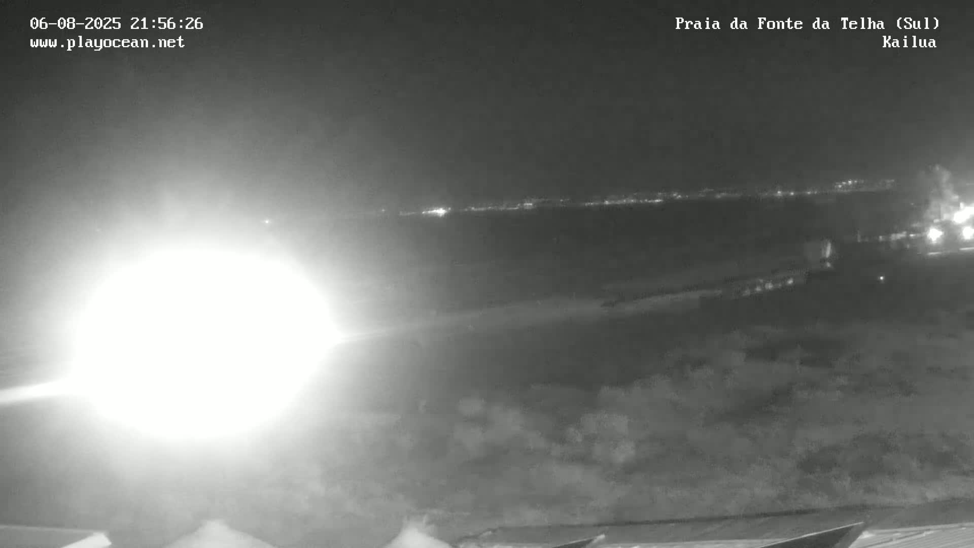 A nighttime, low-light image shows a beach with a bright light source in the foreground and distant city lights on the horizon.