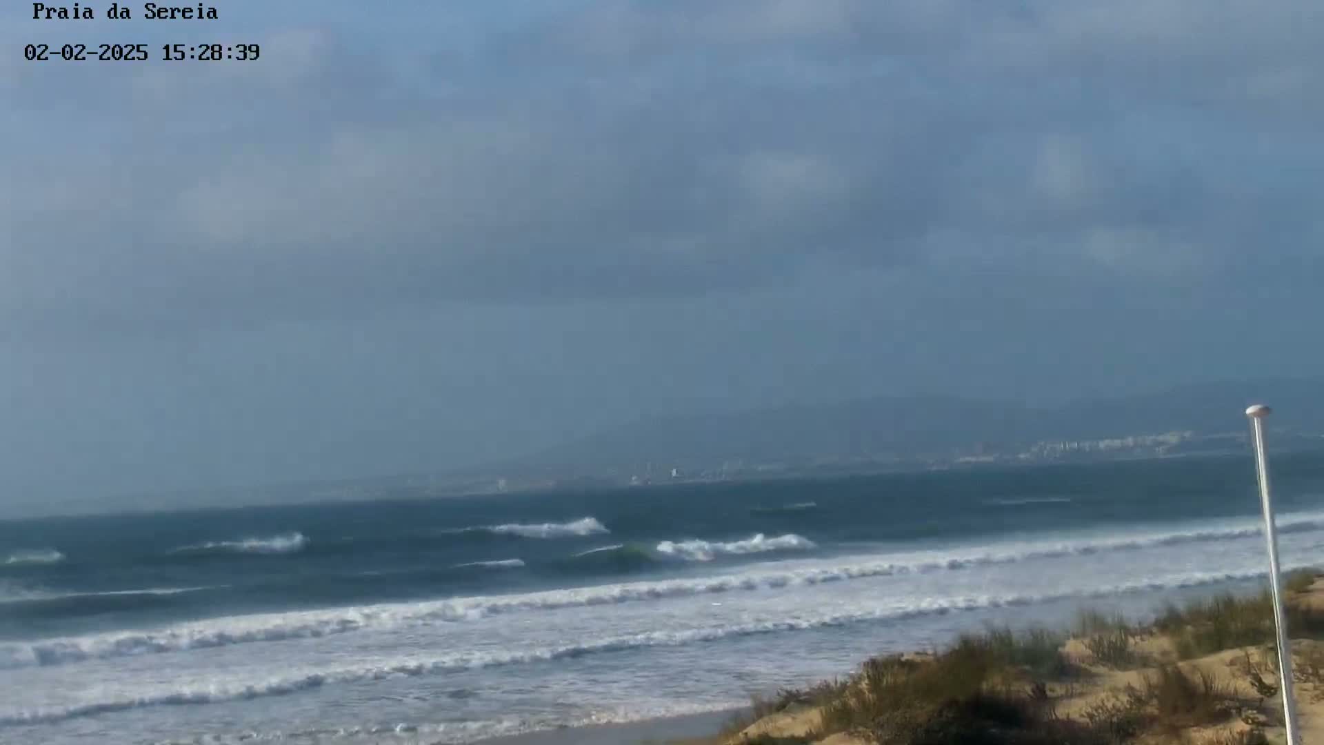 A partly cloudy day shows a view of an ocean beach with moderate waves breaking on the shore, and sand dunes in the foreground.