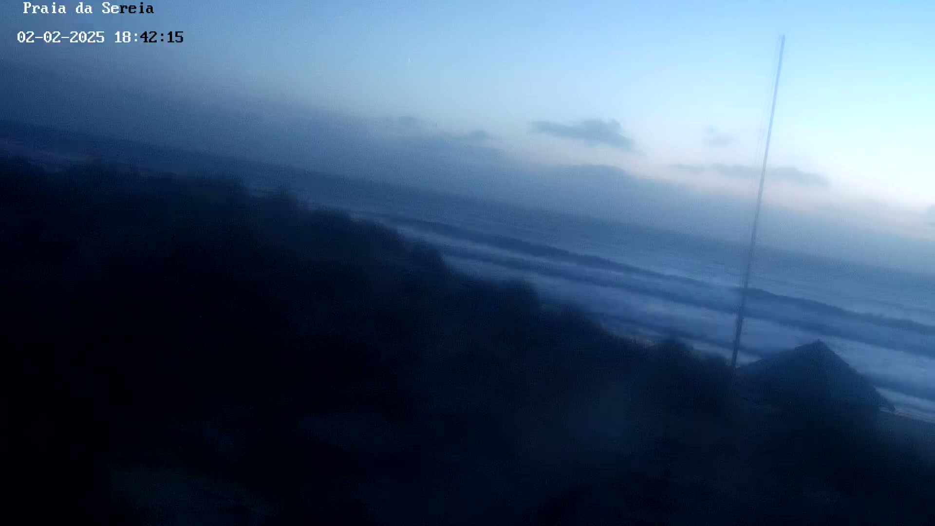 A hazy, twilight view shows a dark, wave-swept ocean with a shadowed shoreline and a tall, thin pole.