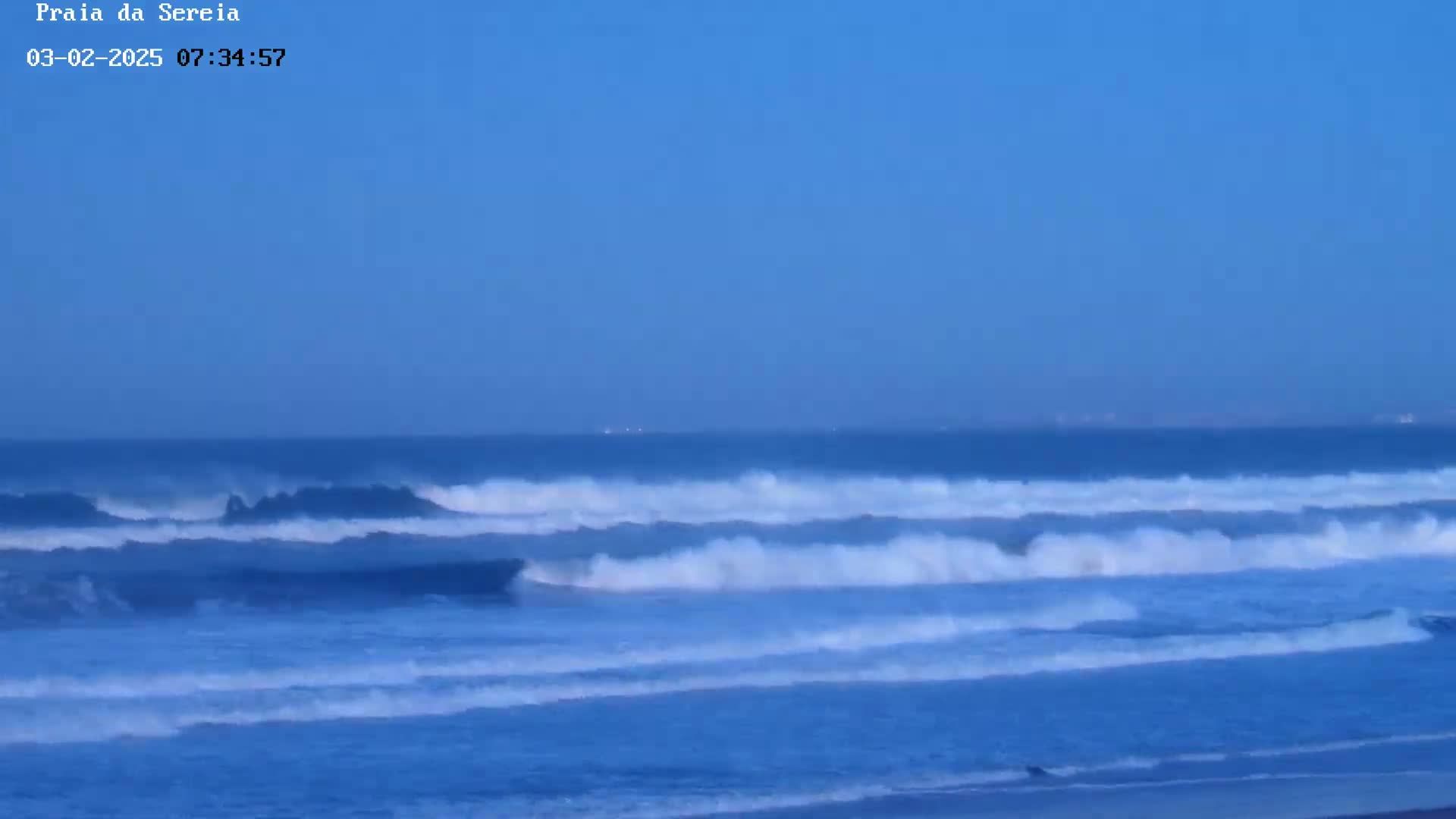 Under a clear, light-blue sky, several sets of ocean waves roll toward a sandy beach.
