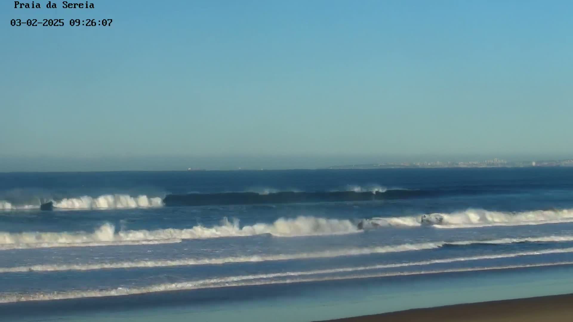 A long expanse of ocean with moderately sized waves crashing on a sandy beach under a clear blue sky.