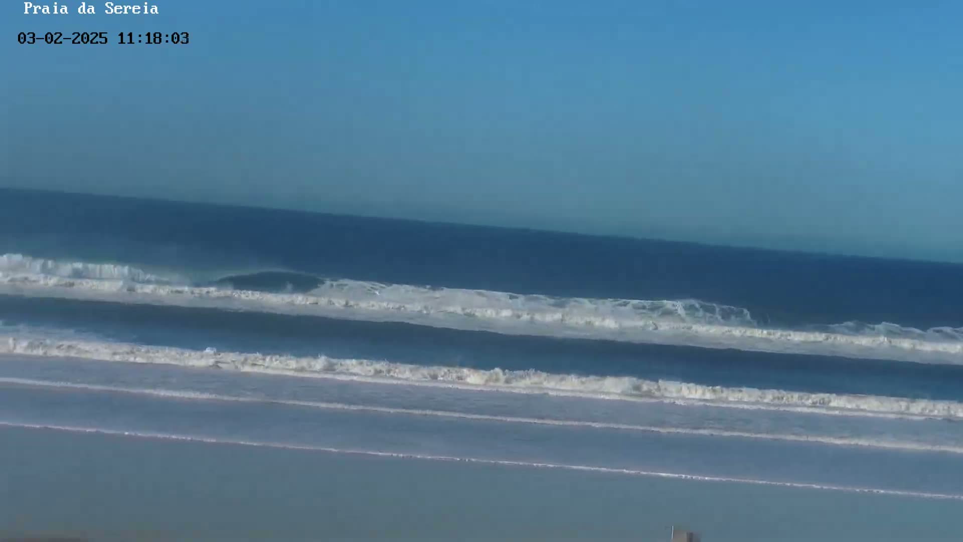 A wide shot shows an ocean beach on a clear day with several sets of waves breaking near the shore.