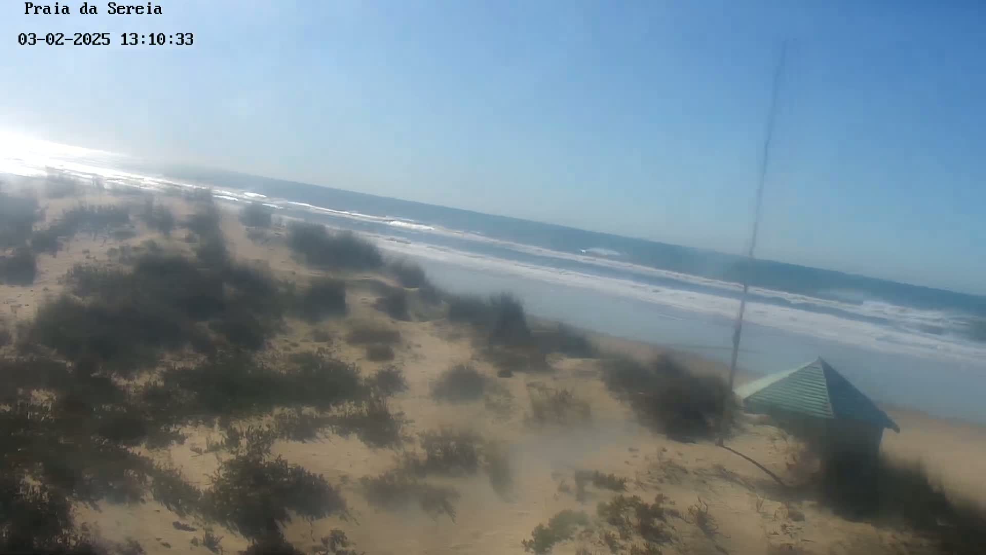 A hazy view shows a sandy beach with sparse vegetation and a small green structure, overlooking a somewhat choppy ocean under a clear sky.