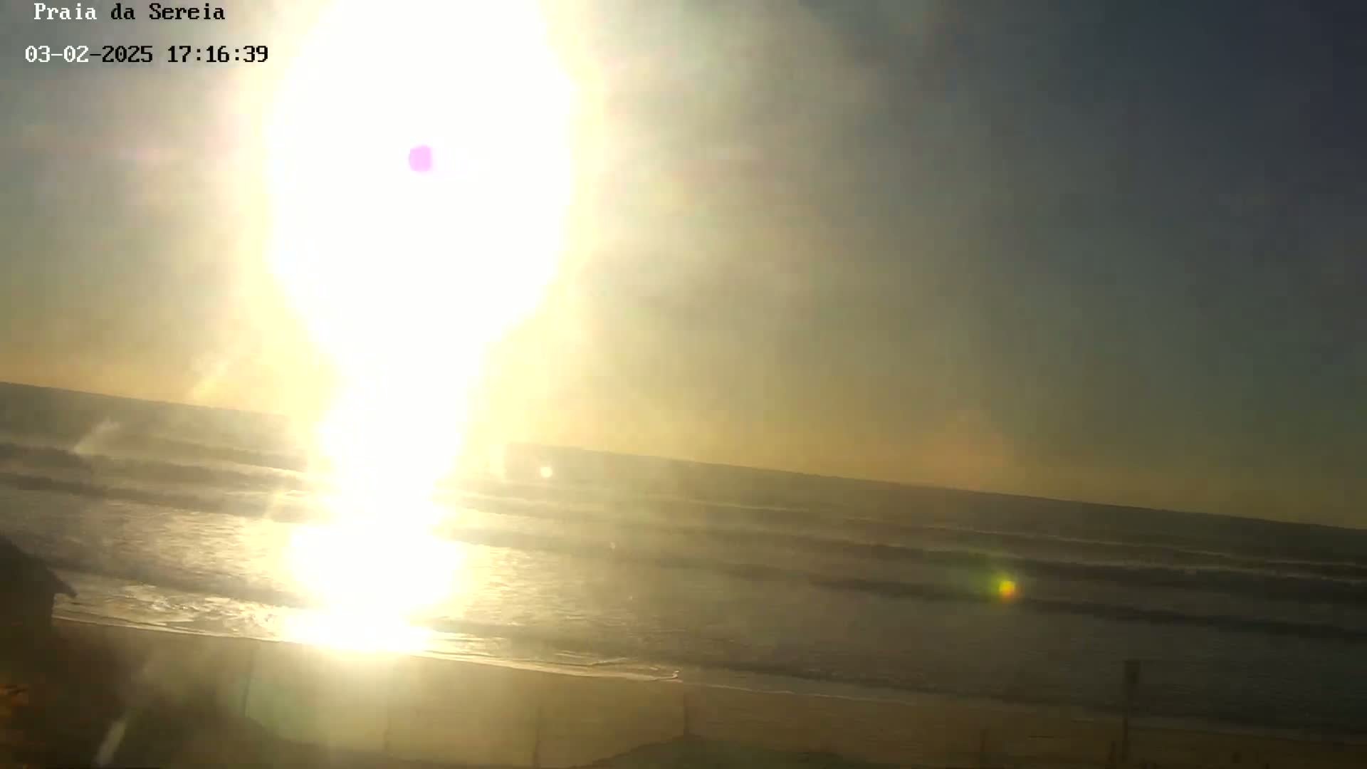A bright, hazy sun shines over a beach with gentle waves rolling onto the shore.