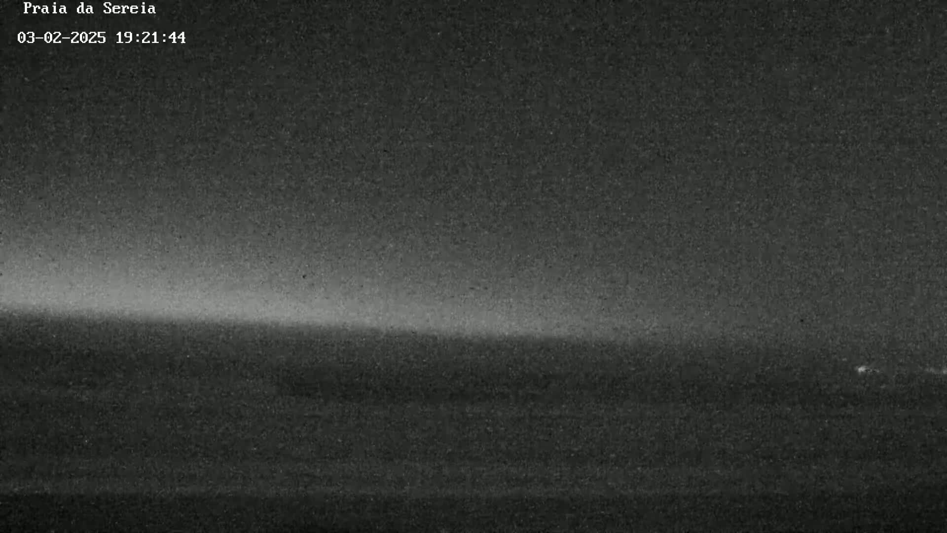 A low-light, grainy image shows a dark, flat landscape, possibly an ocean or beach, under a dark, possibly nighttime sky with a faint, lighter horizon line.