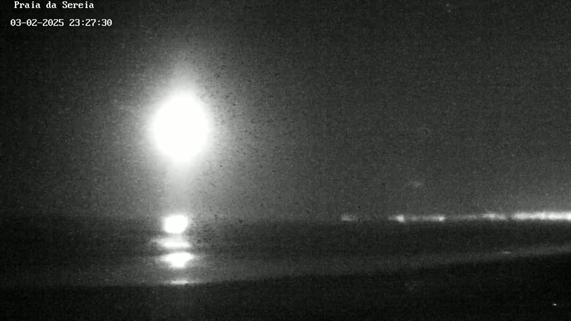 A bright light, possibly the moon, illuminates a dark, nighttime beach scene with a faint horizon line showing distant lights.