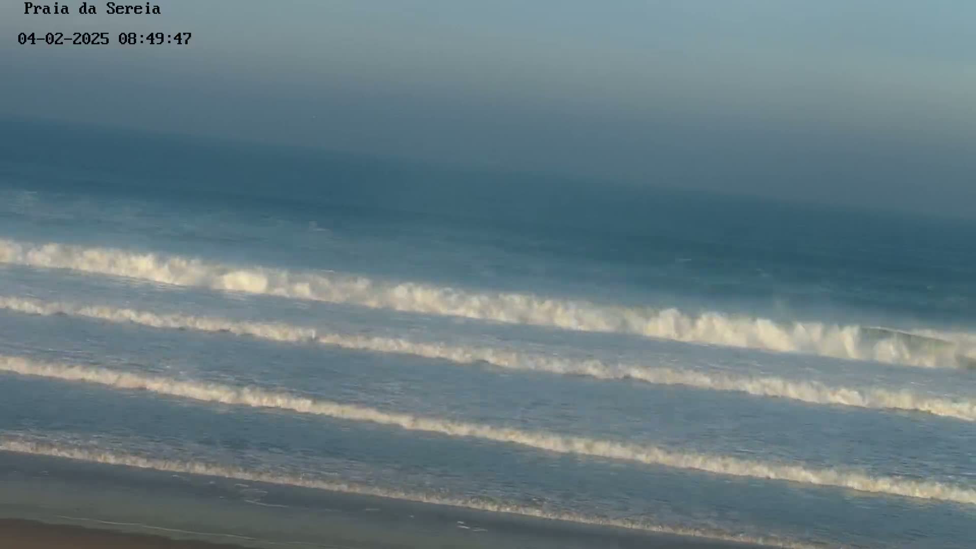 Under a hazy sky, several lines of waves roll onto a sandy beach.