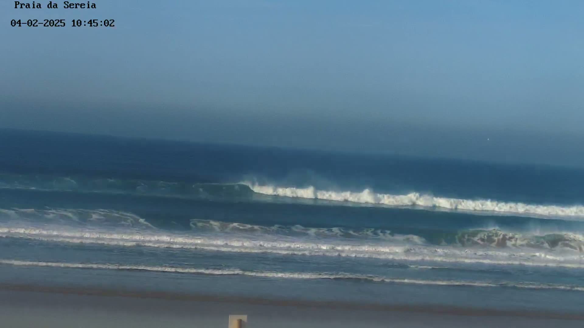 A long, continuous line of waves rolls onto a sandy beach under a clear, light-blue sky.