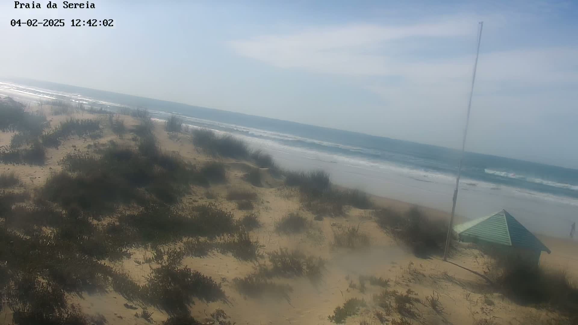 A hazy, sunny day at a sandy beach shows a small, light green structure near the dunes overlooking a wavy ocean.