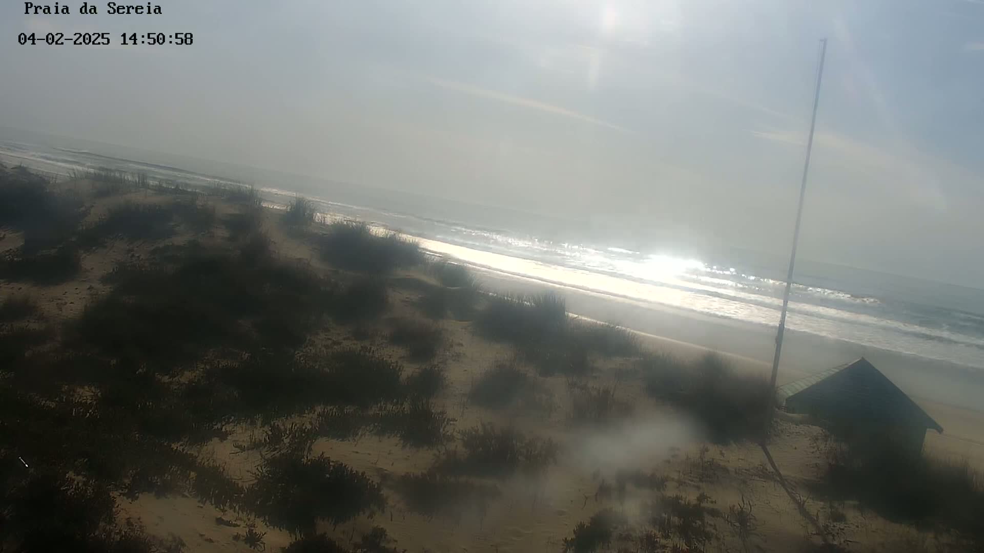 A hazy, sunny day at a sandy beach with low-lying vegetation, gentle waves, and a small structure near the shore.