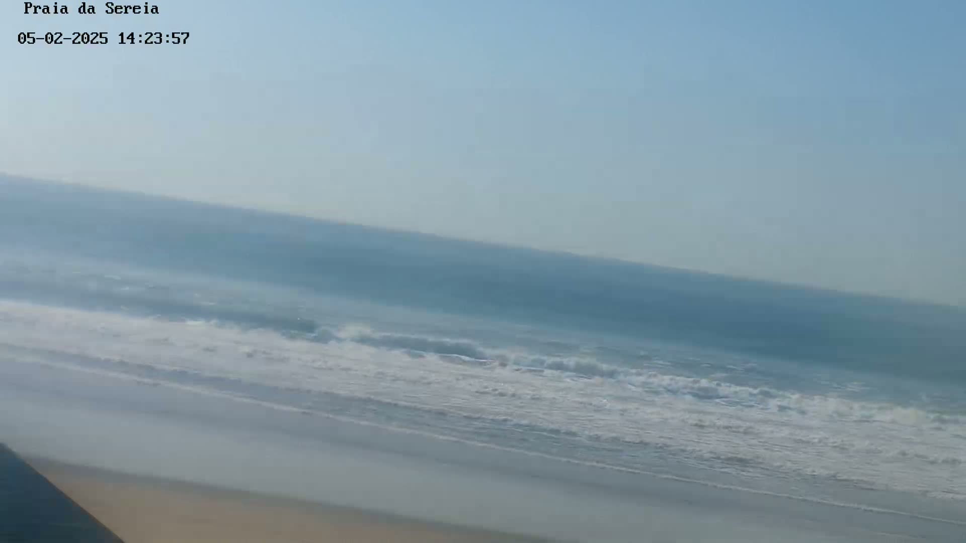 A long expanse of ocean with small waves breaking on a sandy beach under a clear, light blue sky.