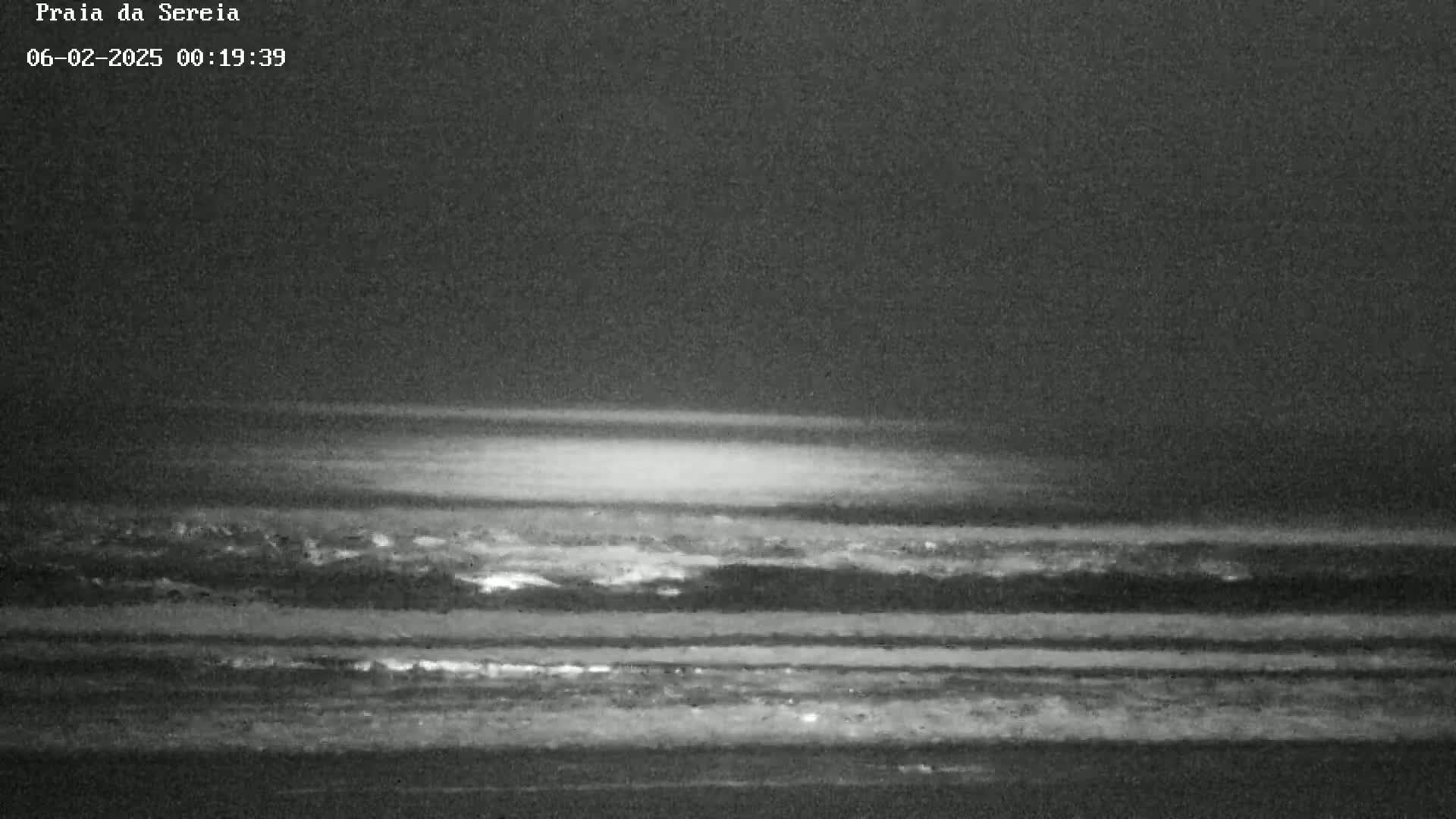 A dark, nighttime beach scene shows a dimly lit ocean with waves gently lapping the shore under a mostly clear sky.