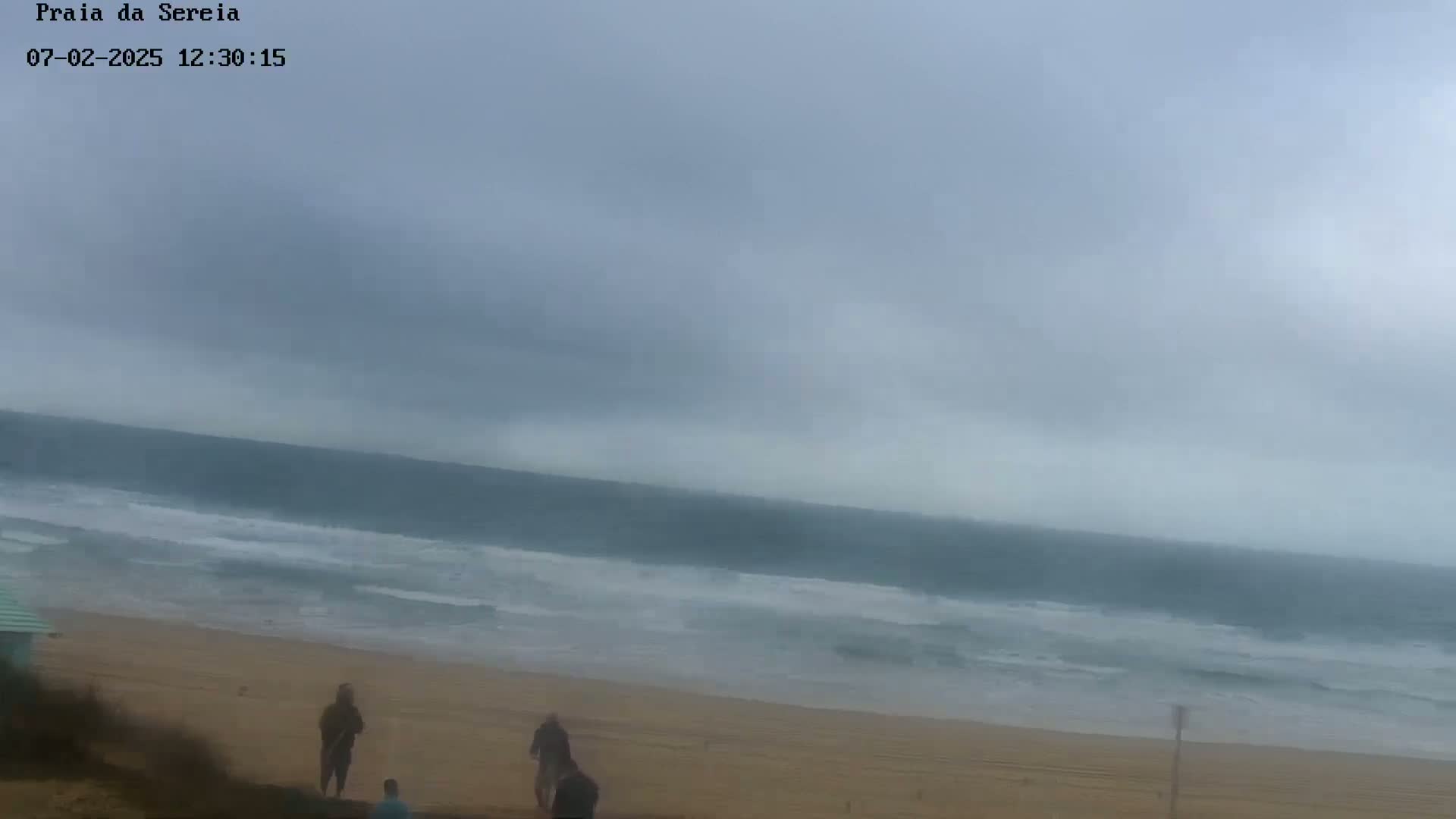 A mostly cloudy, overcast day shows a beach with a few people walking on the sand and waves rolling into the shore.