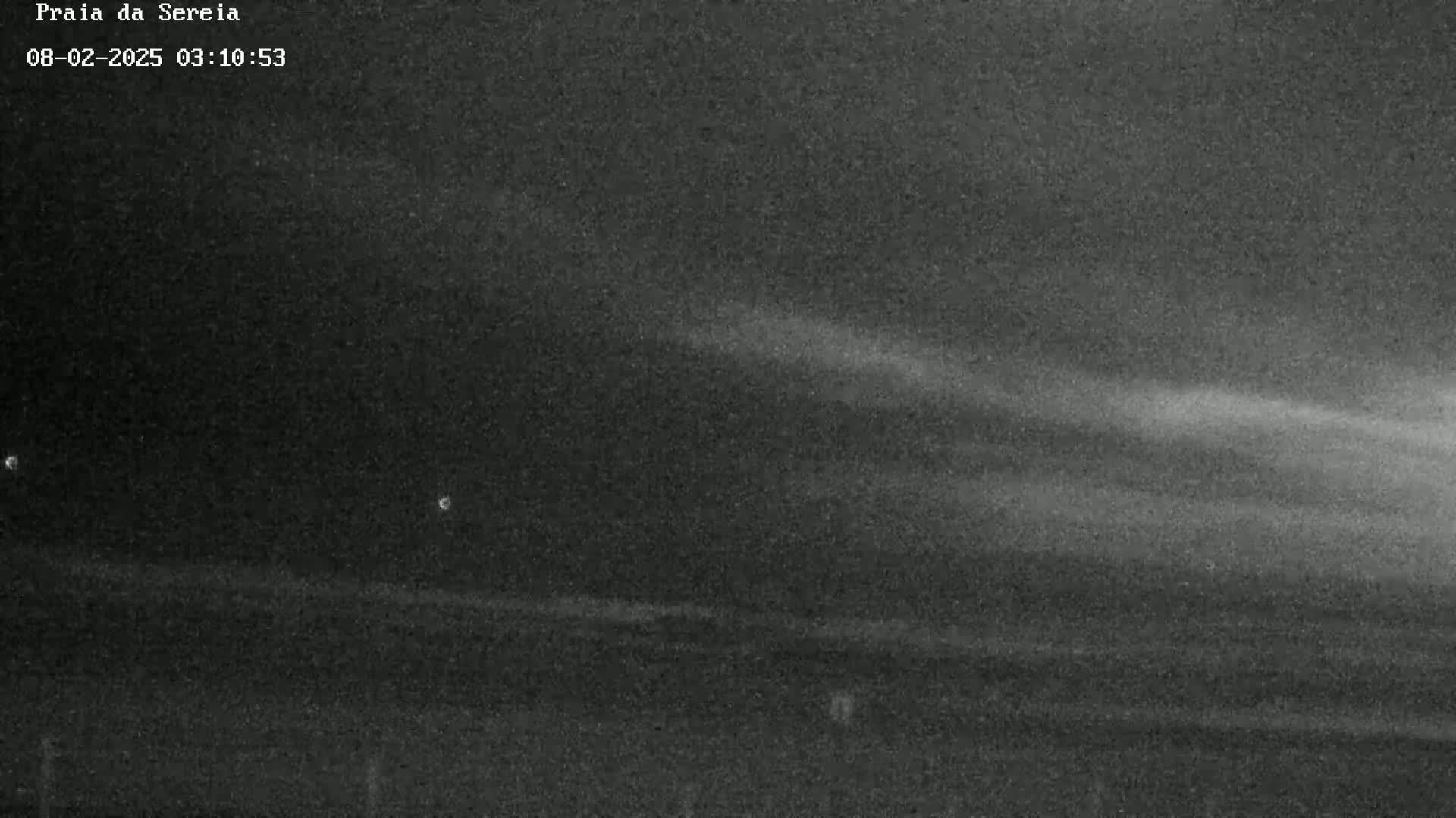 A low-light, nighttime image shows a dark, flat landscape under a mostly clear sky with faint, horizontal streaks of light possibly from a light source.