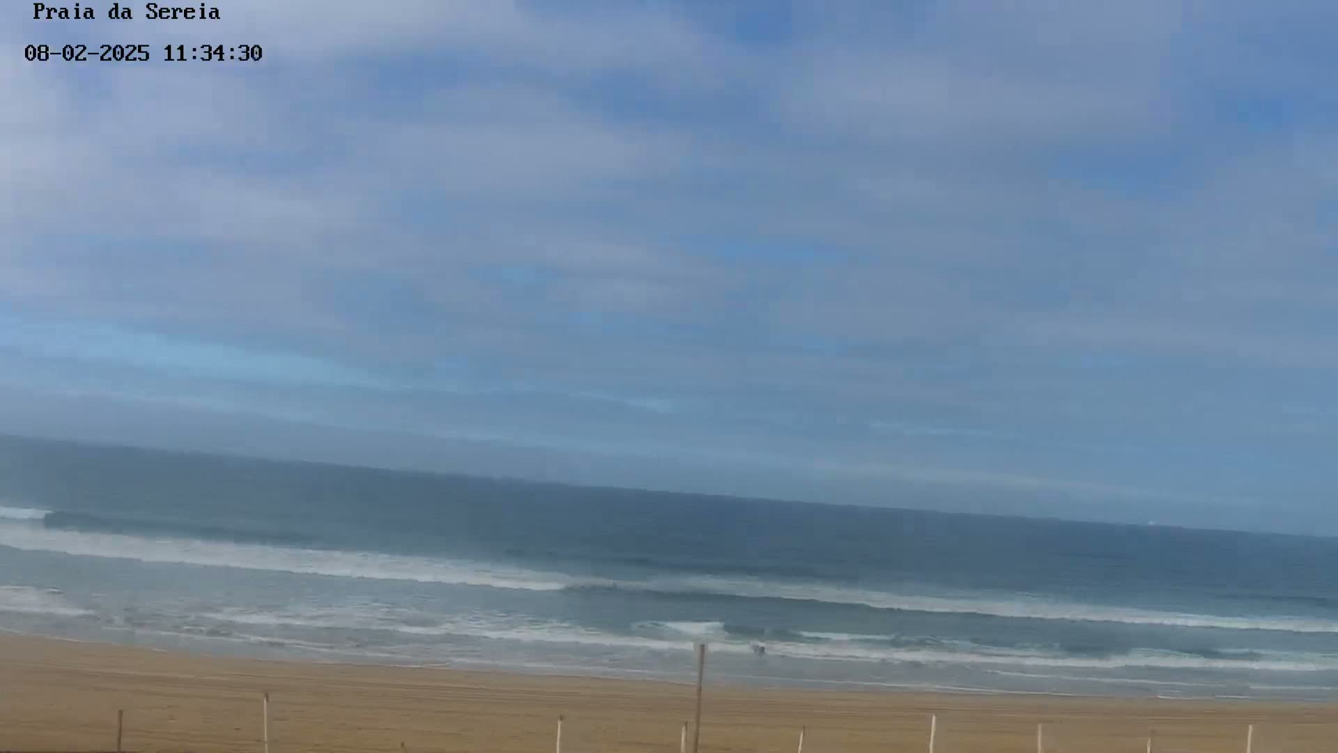 A mostly clear sky overlooks a calm ocean with small waves lapping a sandy beach.