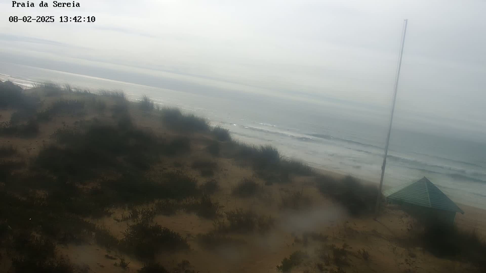 A hazy view of a sandy beach with low-lying vegetation, gentle waves, and a small green structure, all under overcast skies.