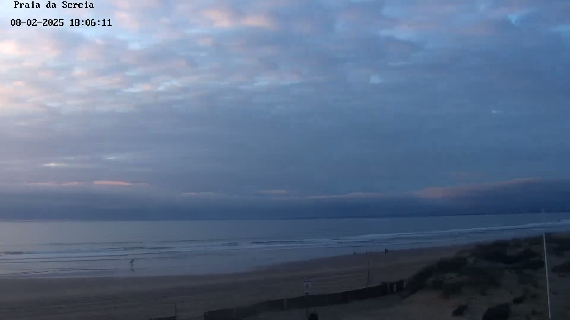 A mostly cloudy, twilight sky overlooks a calm ocean and sandy beach with a few people visible in the distance.