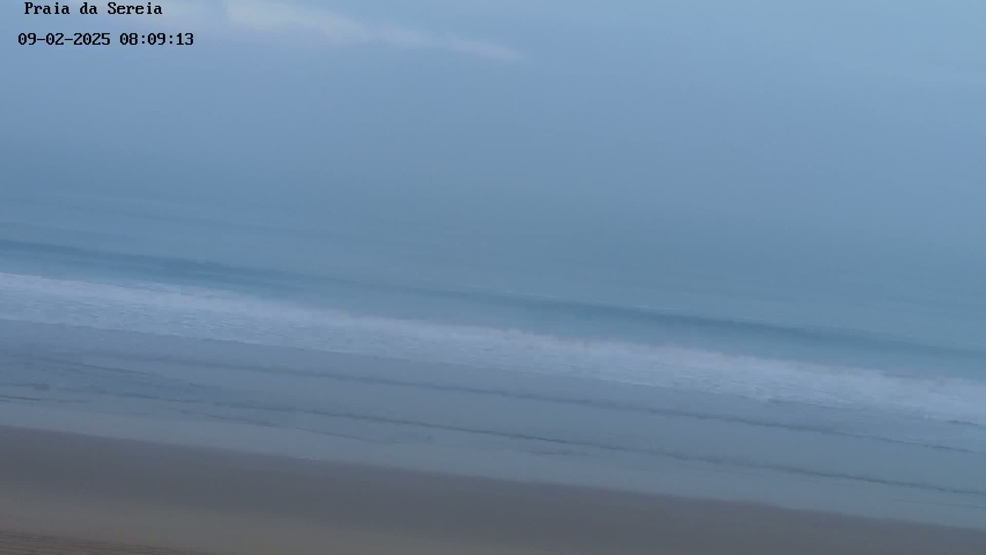 A hazy, overcast sky overlooks a calm ocean with gentle waves lapping onto a sandy beach.