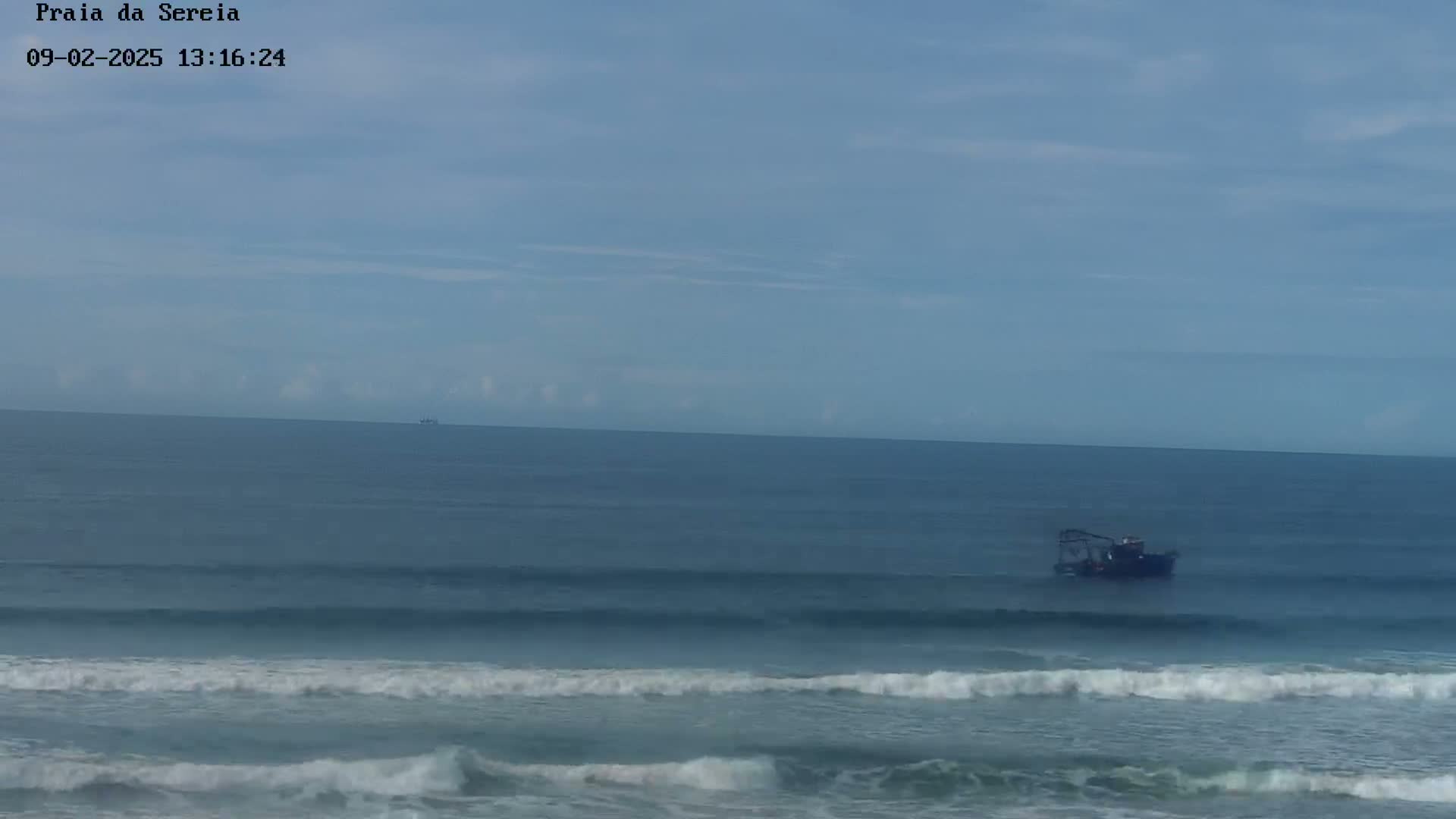 A small boat sits on the ocean under a mostly clear sky, with gentle waves rolling onto the shore.