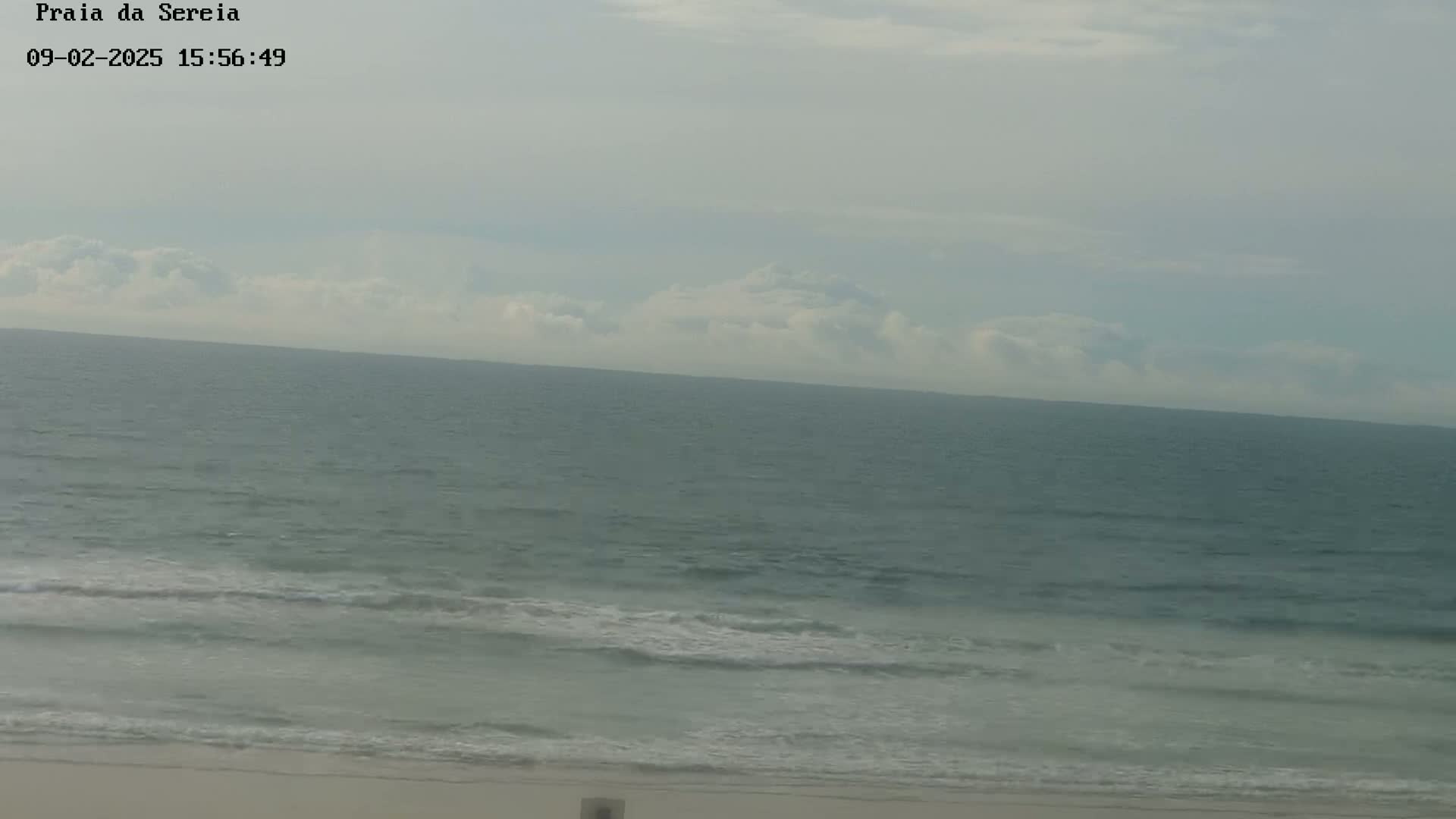 A mostly cloudy day shows a calm ocean meeting a sandy beach with small waves gently lapping the shore.