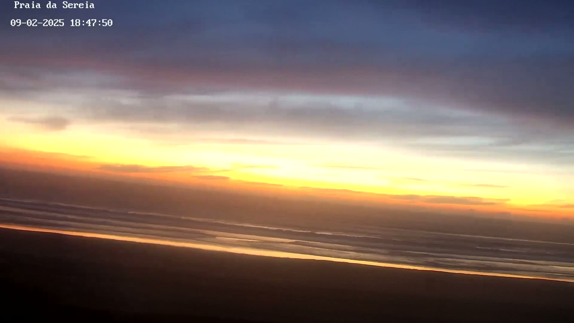 A beach scene at sunset shows a wide expanse of ocean with small waves under a colorful sky of yellow, orange, and purple hues.