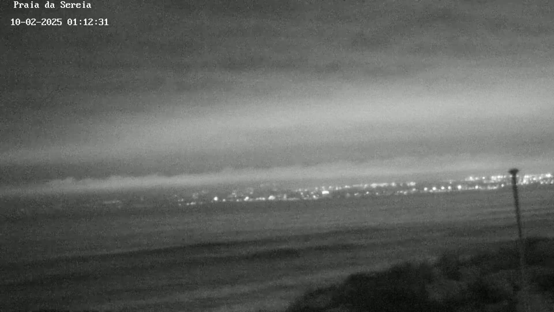A nighttime coastal scene shows a dimly lit city in the distance across a dark body of water, under a cloudy sky.