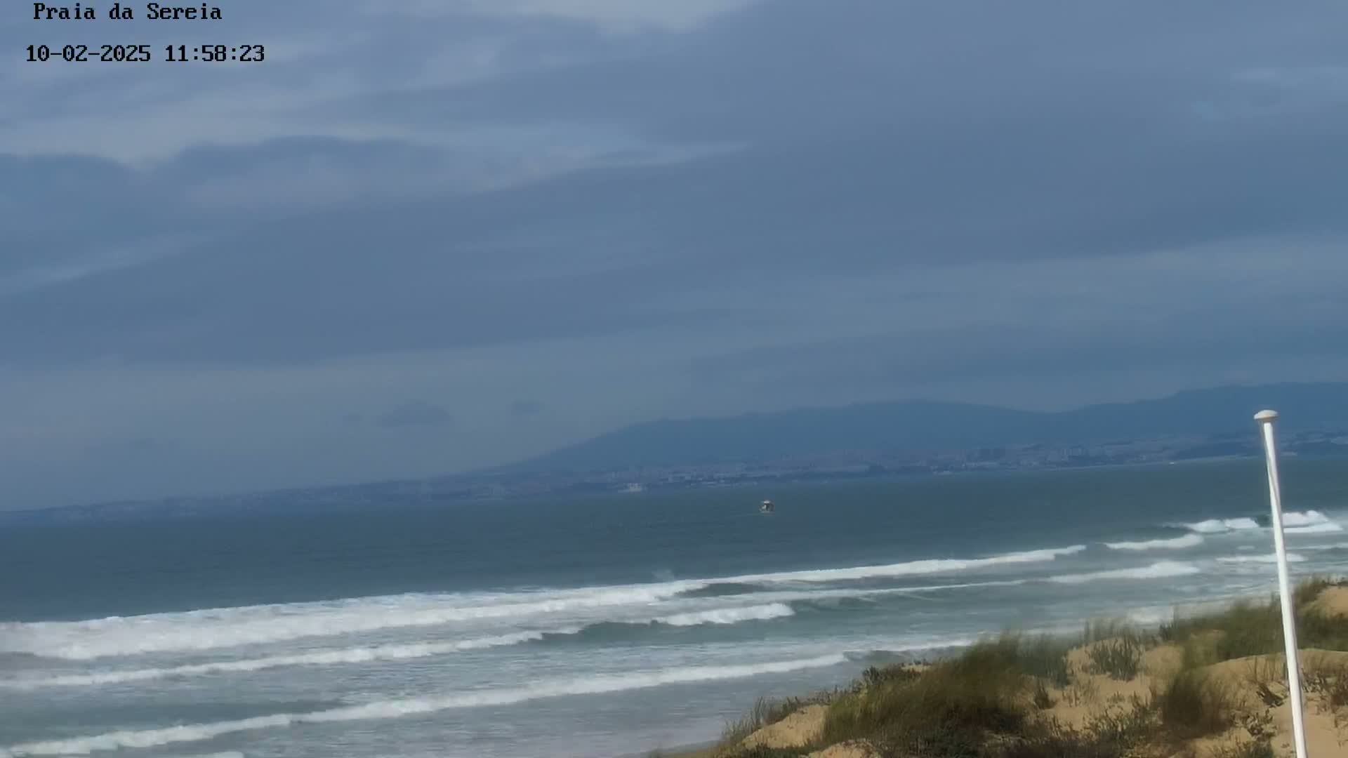 Under an overcast sky, ocean waves roll onto a sandy beach with dunes and sparse vegetation in the foreground, and distant mountains visible on the horizon.