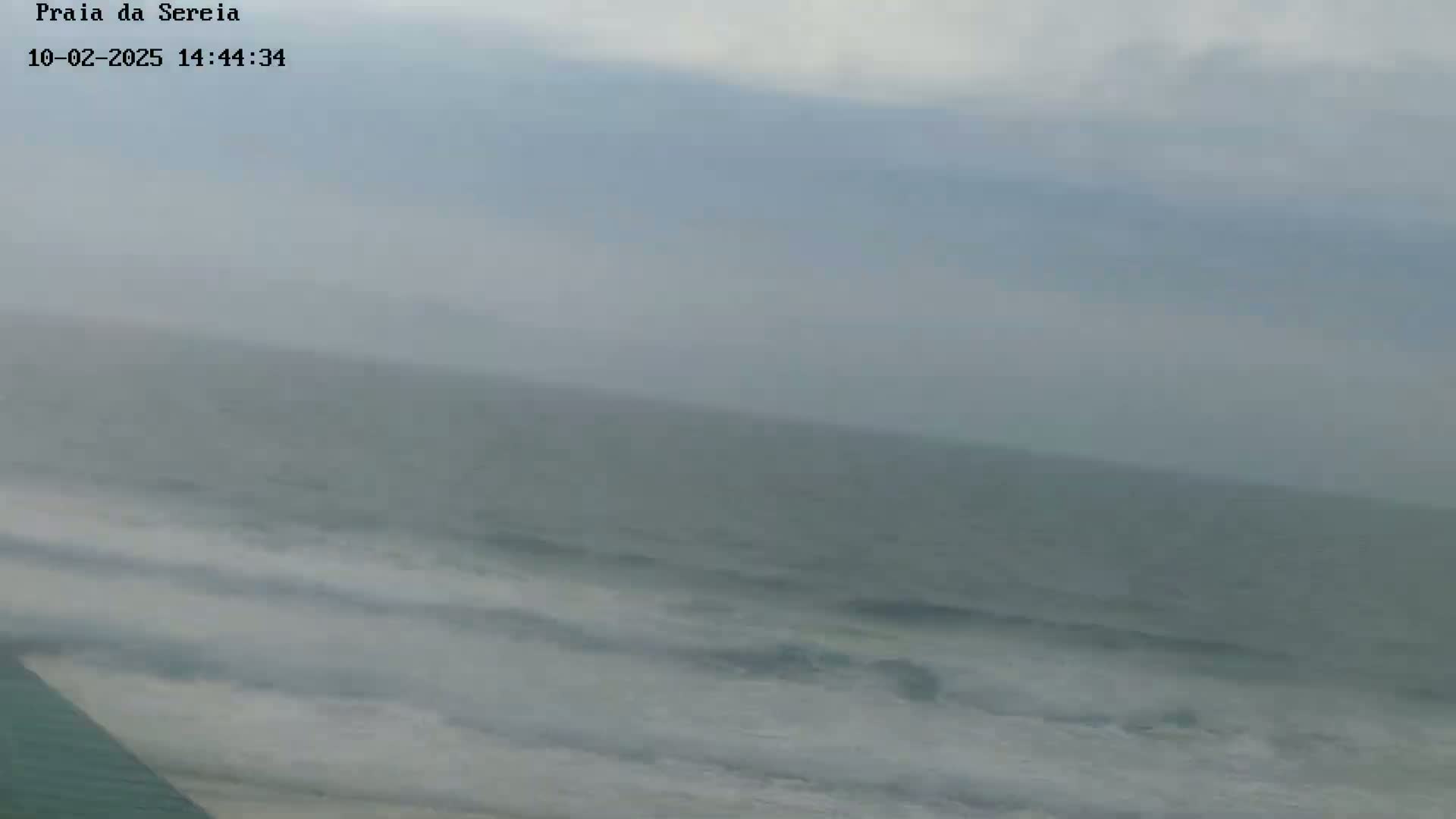 A hazy overcast sky looms over a slightly choppy ocean with waves breaking on a beach.