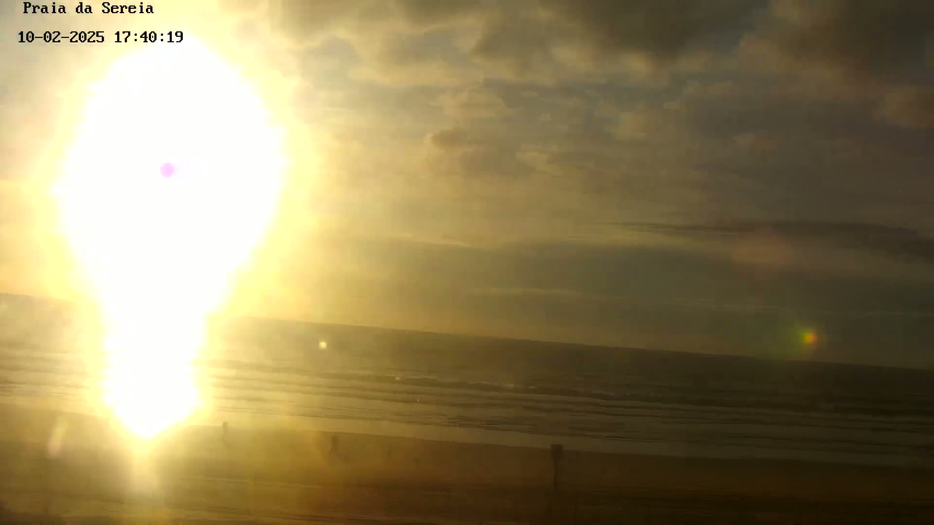 A bright, hazy sunset over a calm ocean beach with a few people silhouetted on the sand.