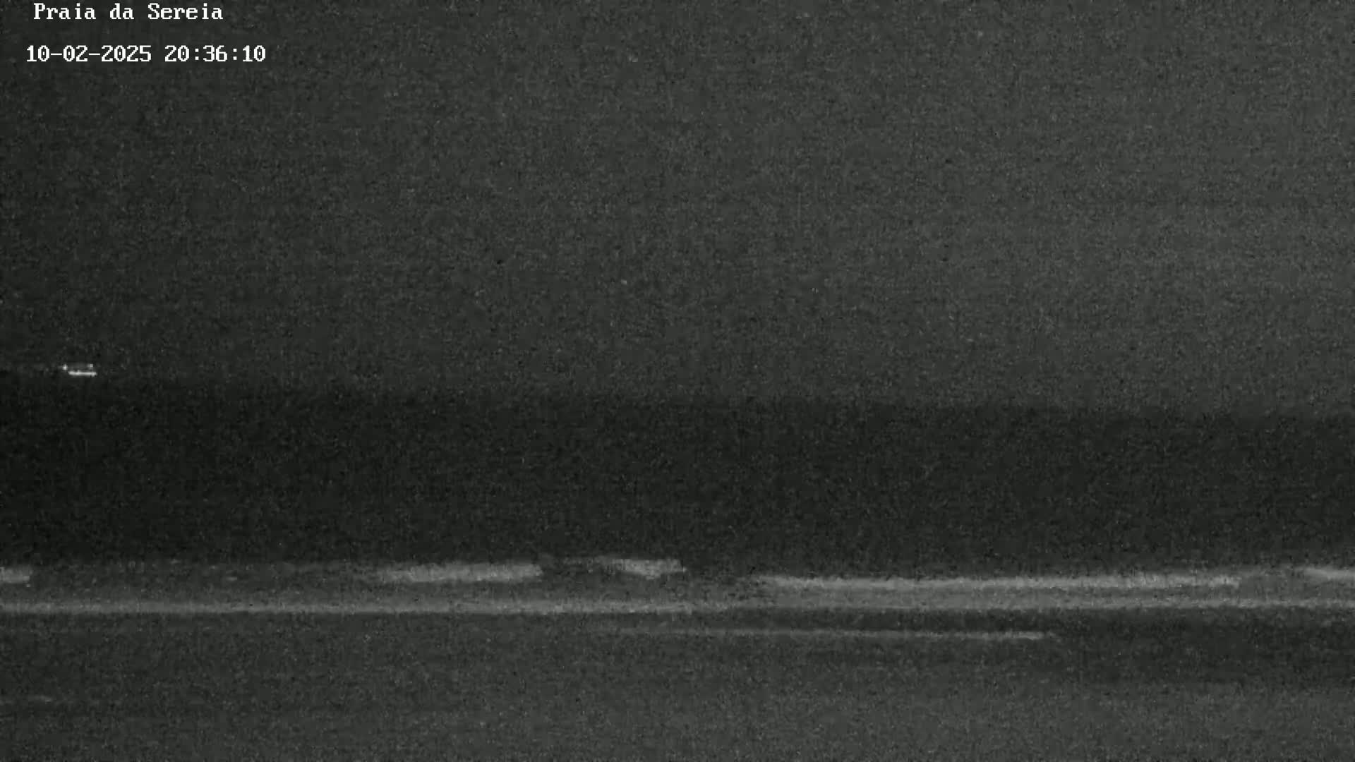 A nighttime, low-light image shows a dark beach with a faint line of waves and a dark horizon under a dark sky.