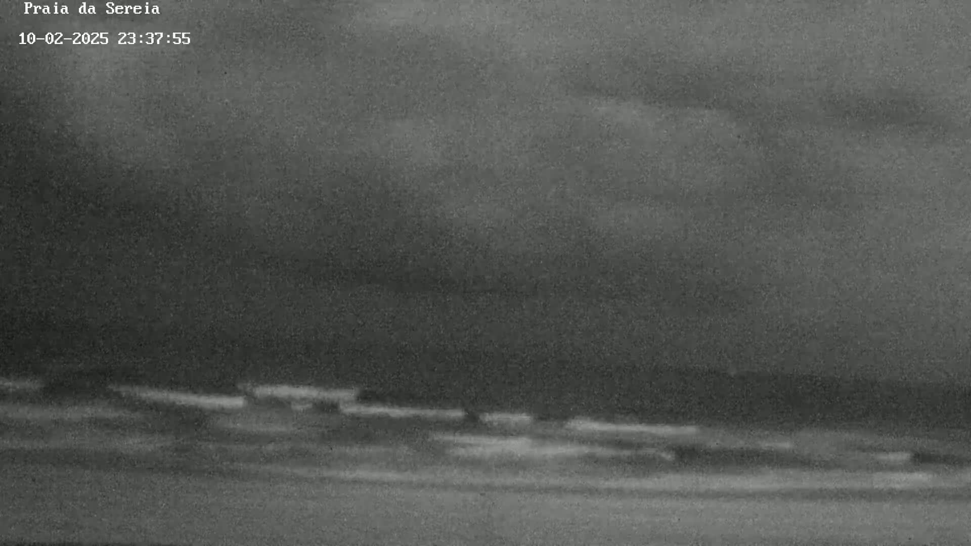 A nighttime, overcast beach scene shows dark, wavy water meeting a dark shoreline under a cloudy sky.