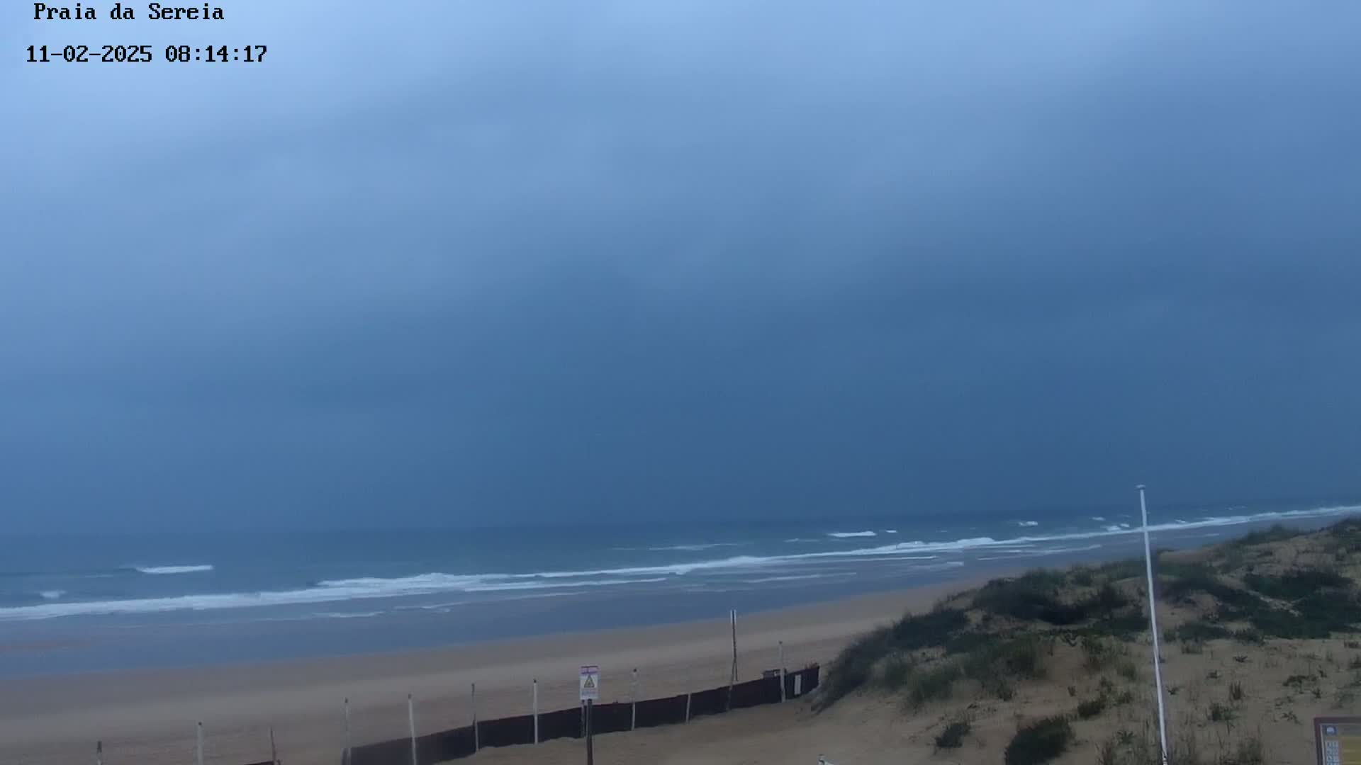A mostly cloudy sky hangs over a sandy beach with small waves lapping the shore, and dunes in the foreground.