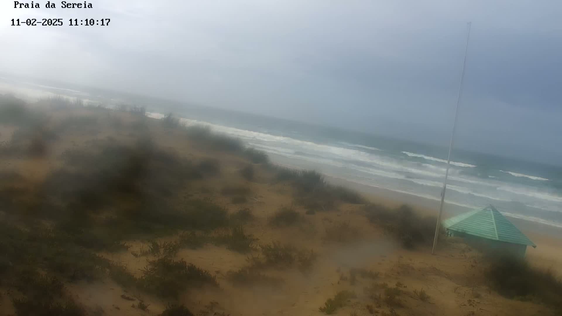 A hazy, overcast day at a sandy beach shows a small green structure near the shore, with waves breaking in the ocean.