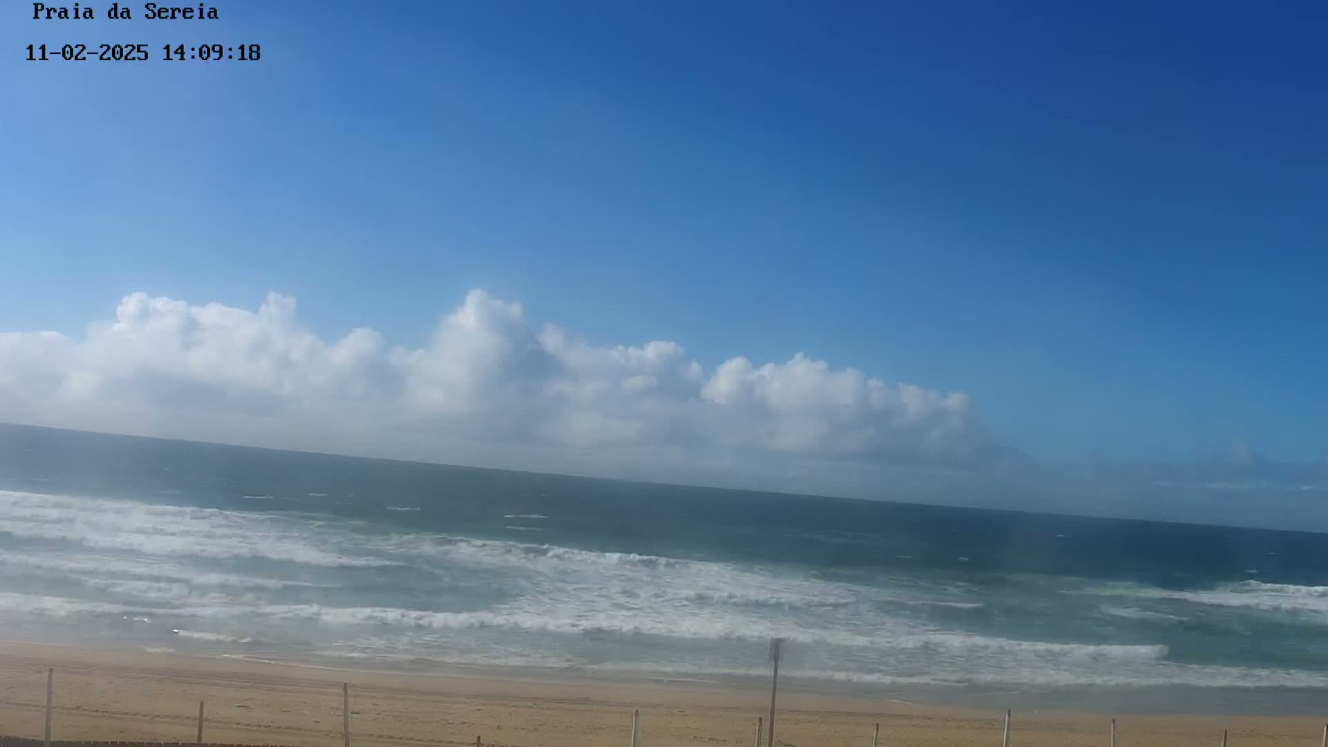 A sandy beach with a fence stretches along a coastline, where ocean waves roll in under a partly cloudy, bright blue sky.